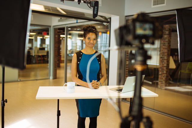 Jourdan facing cameras in studio standing behind a table with a computer and coffee mug with camer